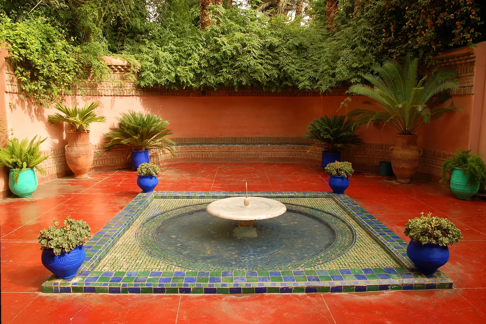 A terracotta-red courtyard with a round tiled fountain in blue and green mosaic, surrounded by blue and green flowerpots and a low wall of greenery