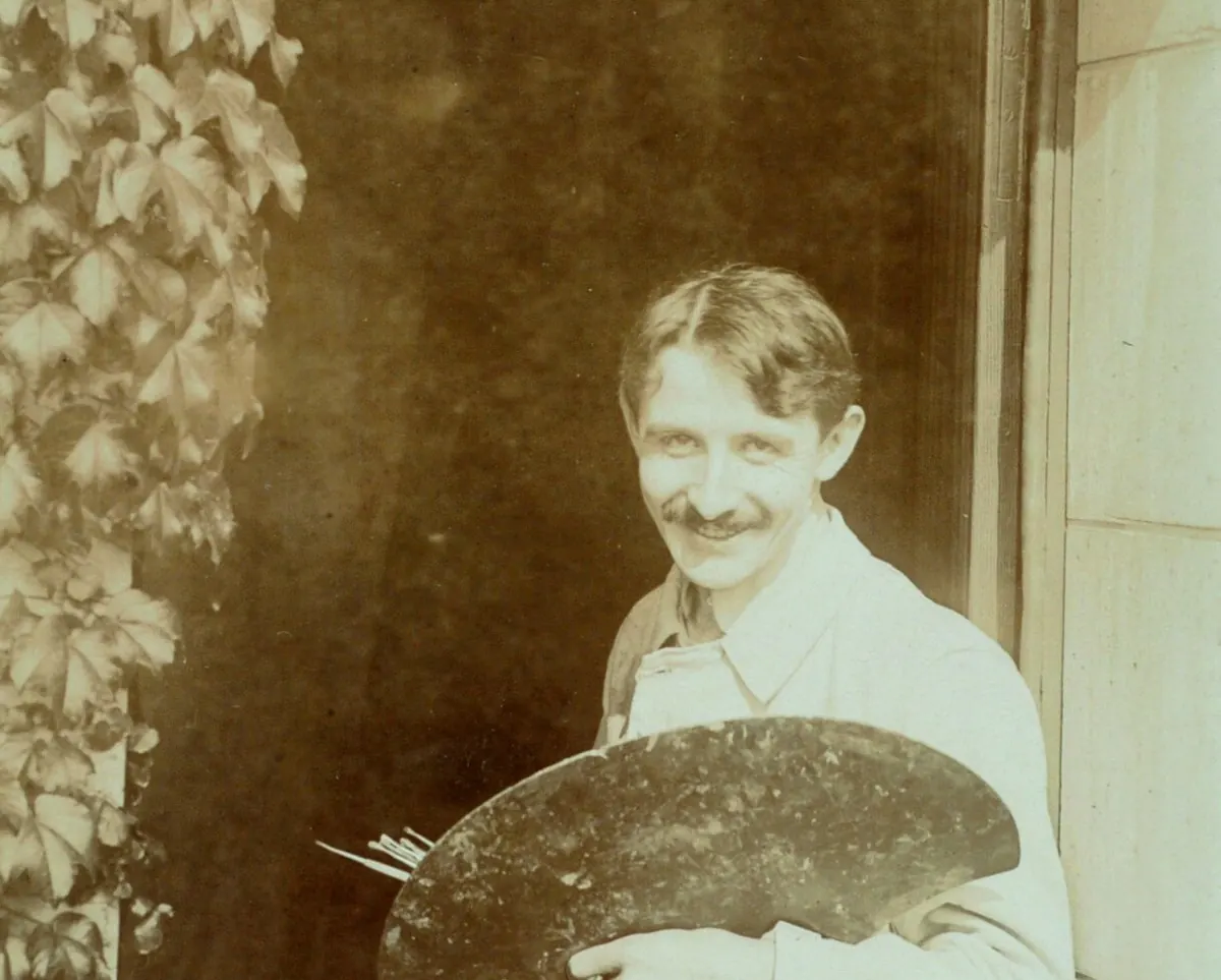 Jacques Majorelle smiling in a doorway under an ivy-covered arch, holding a painter's palette, in a sepia photograph from around 1910